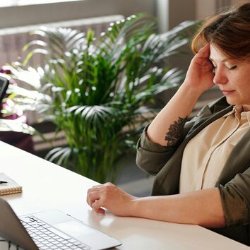 Frau greift sich an Schläfe, sie sitzt vor einem PC